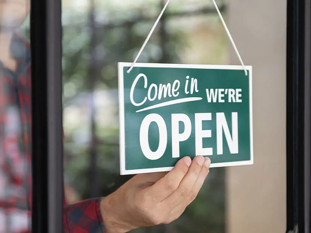 someone on the other side of a widow holding a sign saying "come in we're open"