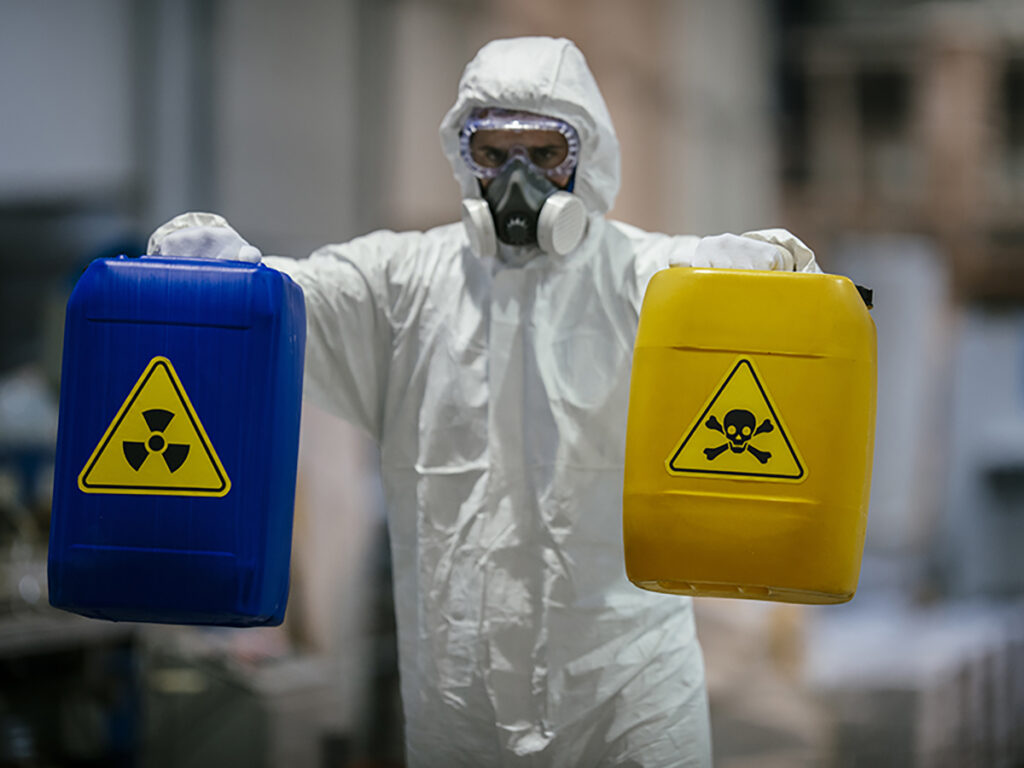 worker in PPE, a hazmat suit, goggles, and air mask respirator carrying 2 plastic containers of hazardous chemicals
